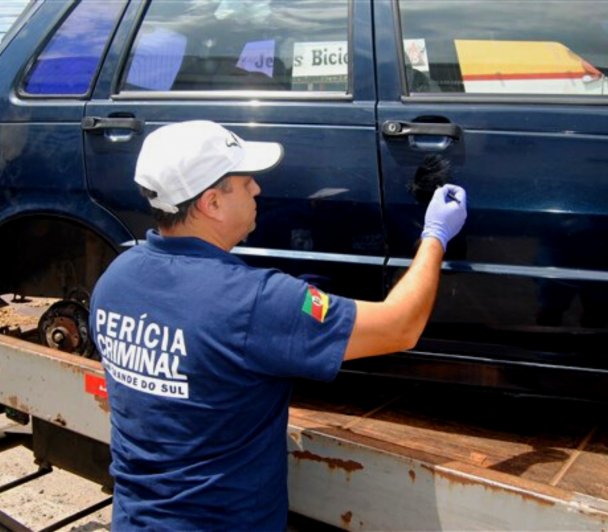 Homem com roupa azul escuro da perícia criminal do Rio Grande do Sul e bone branco realizando a perícia no carro azul marinho