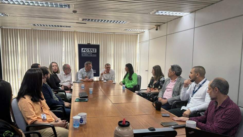Foto mostra reunião na sala da Fgtas, com pessoas sentadas a volta de uma mesa de trabalho. O banner da Fgtas está atrás dos secretários.